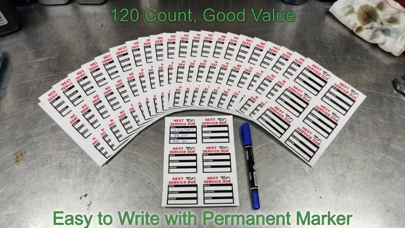 The image showcases a large pack of oil change reminder stickers arranged in a semi-circular fan pattern on a textured metallic surface, resembling a garage workbench. In the front, a single sheet of stickers and a blue permanent marker are visible.